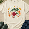 An off-white graphic t-shirt laid flat on a textured beige background, styled with green leaves, black rain boots, and blue jeans. The t-shirt features a retro-style illustration of a red beet with green leaves and orange and yellow graphic elements, with the text 'DON'T STOP THE BEET' printed above it.