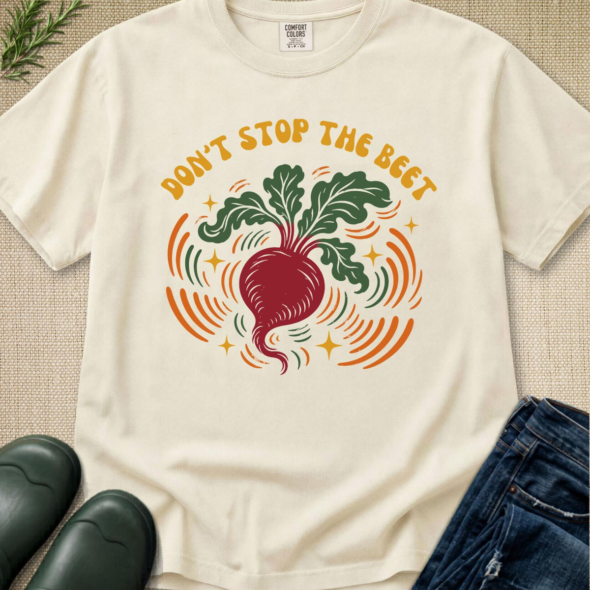 An off-white graphic t-shirt laid flat on a textured beige background, styled with green leaves, black rain boots, and blue jeans. The t-shirt features a retro-style illustration of a red beet with green leaves and orange and yellow graphic elements, with the text 'DON'T STOP THE BEET' printed above it.