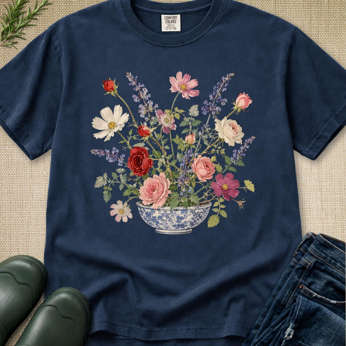 A navy blue short-sleeve t-shirt with a colorful floral print of a flower arrangement in a blue and white patterned bowl, styled in a flat lay with jeans, green rain boots, and a burlap background.