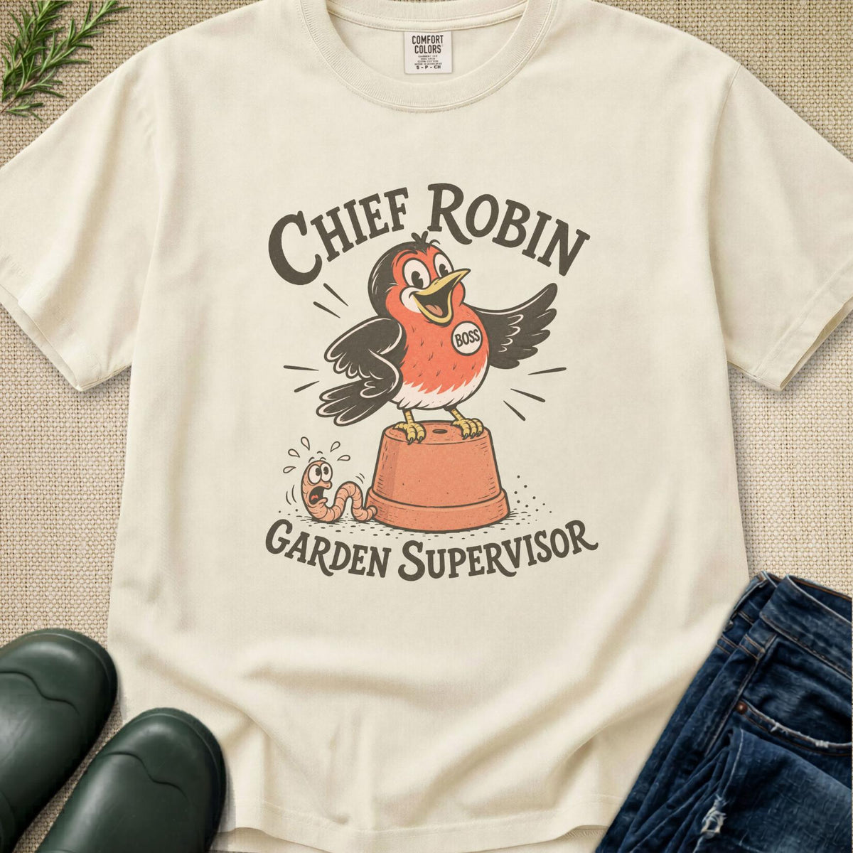 An off-white graphic t-shirt laid flat on a textured beige background, styled with dark green rain boots, blue jeans, and a sprig of greenery. The t-shirt features a cartoon graphic of a robin perched on a terracotta pot with the text 'CHIEF ROBIN GARDEN SUPERVISOR'.