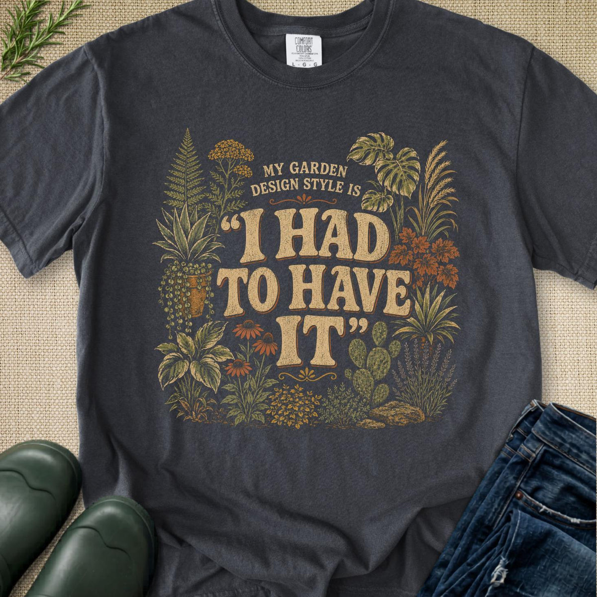 A dark charcoal gray graphic t-shirt laid flat on a textured beige burlap background, styled with green rain boots, blue jeans, and a small sprig of greenery. The t-shirt features a vintage-style botanical illustration of plants and cacti surrounding the text 'MY GARDEN DESIGN STYLE IS "I HAD TO HAVE IT"'.