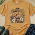 A tan graphic t-shirt laid flat on a textured beige background, styled with green leaves, black rain boots, and blue jeans. The t-shirt features a central graphic of a robin bird perched on a garden tool surrounded by wildflowers and the text 'EVERY SEASON SAME FRIEND'.