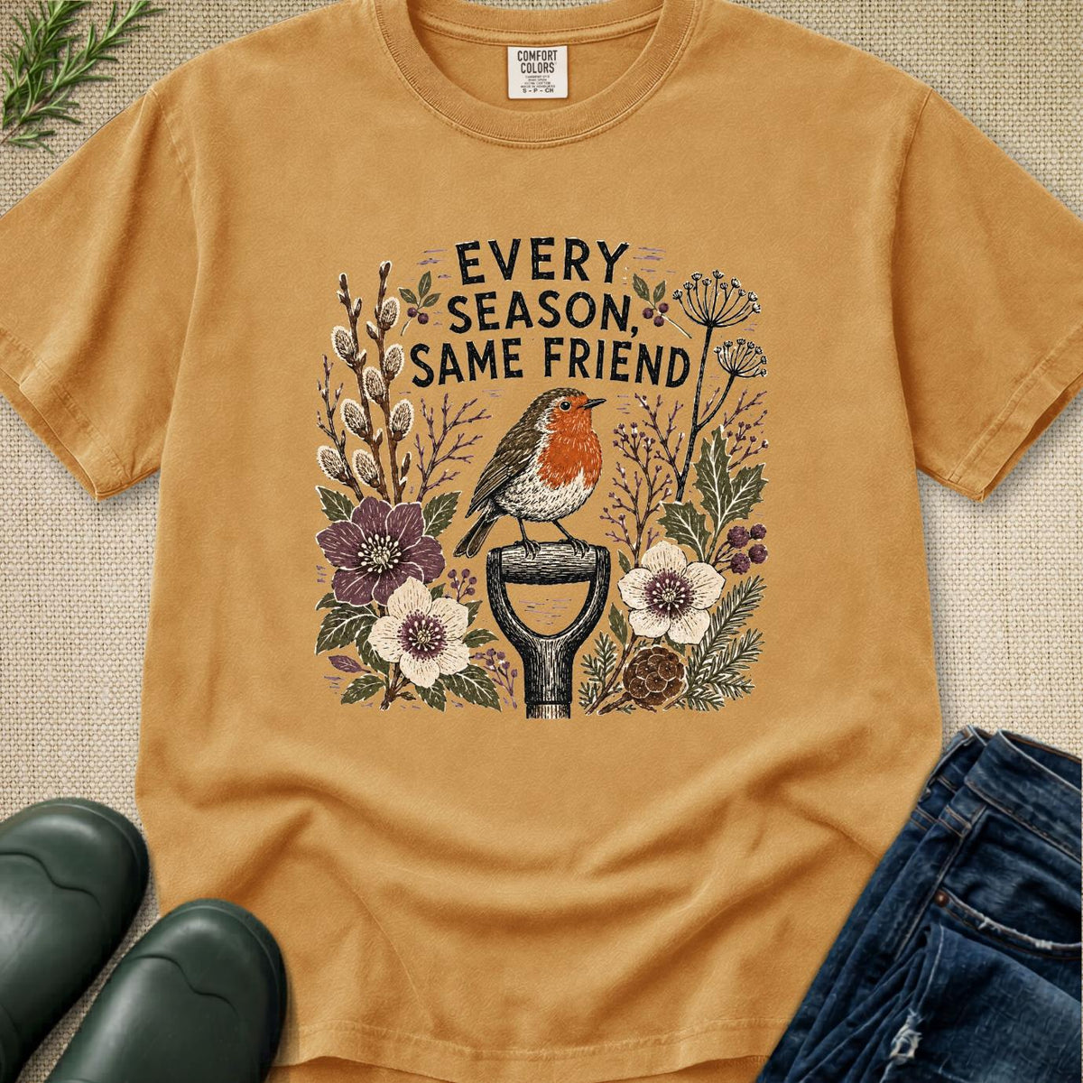 A tan graphic t-shirt laid flat on a textured beige background, styled with green leaves, black rain boots, and blue jeans. The t-shirt features a central graphic of a robin bird perched on a garden tool surrounded by wildflowers and the text 'EVERY SEASON SAME FRIEND'.