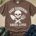A brown short-sleeve graphic T-shirt laid flat on a textured beige surface, with white distressed text reading 'RAGE GARDENING SAVES LIVES' around a skull and crossed gardening tools. Partial jeans, green boots, and a sprig of leaves are placed around the shirt as styling props.