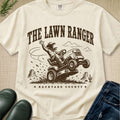 An off-white graphic t-shirt featuring a brown illustration of a cowboy riding a lawnmower with the text 'THE LAWN RANGER' and 'BACKYARD COUNTY'. The shirt is styled in a flat-lay arrangement with blue jeans, green rain boots, and a small plant on a textured beige background.
