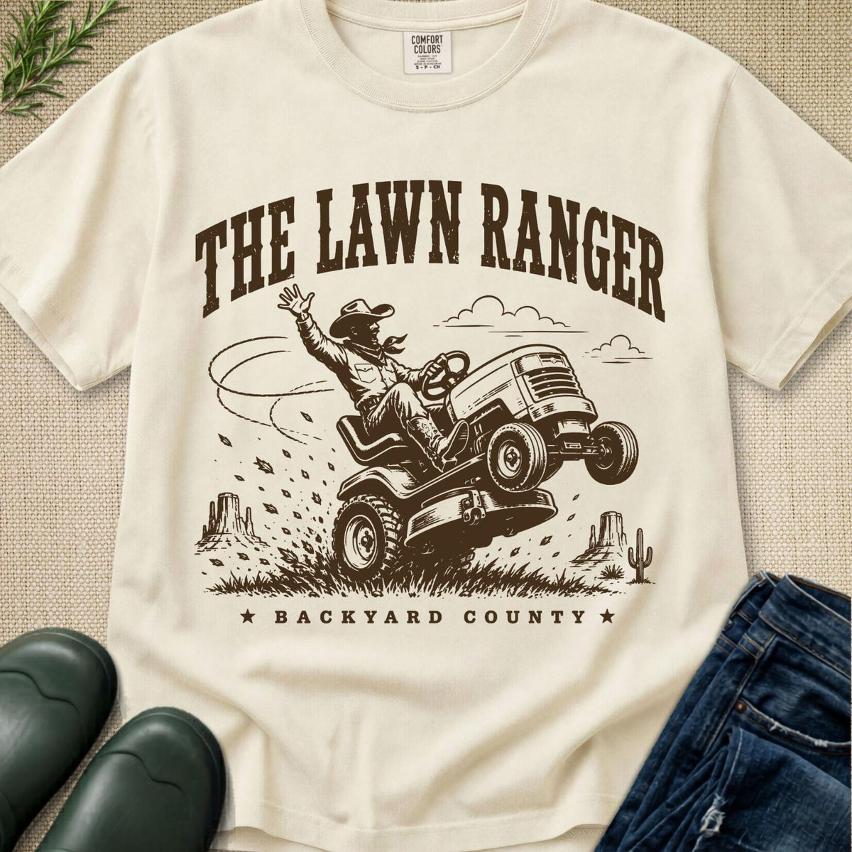 An off-white graphic t-shirt featuring a brown illustration of a cowboy riding a lawnmower with the text 'THE LAWN RANGER' and 'BACKYARD COUNTY'. The shirt is styled in a flat-lay arrangement with blue jeans, green rain boots, and a small plant on a textured beige background.