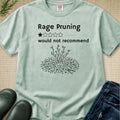A light sage green graphic t-shirt laid flat on a beige textured background, styled with black rubber boots and blue jeans. The t-shirt features a black graphic of a pruned branch and the text 'Rage Pruning would not recommend' with a one-star rating.