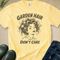 A pale yellow graphic T-shirt laid flat on a textured beige surface, with dark brown text reading 'GARDEN HAIR DON'T CARE' and an illustrated woman with curly hair. Jeans, green rain boots, and a small sprig of greenery are placed around it as styling props.