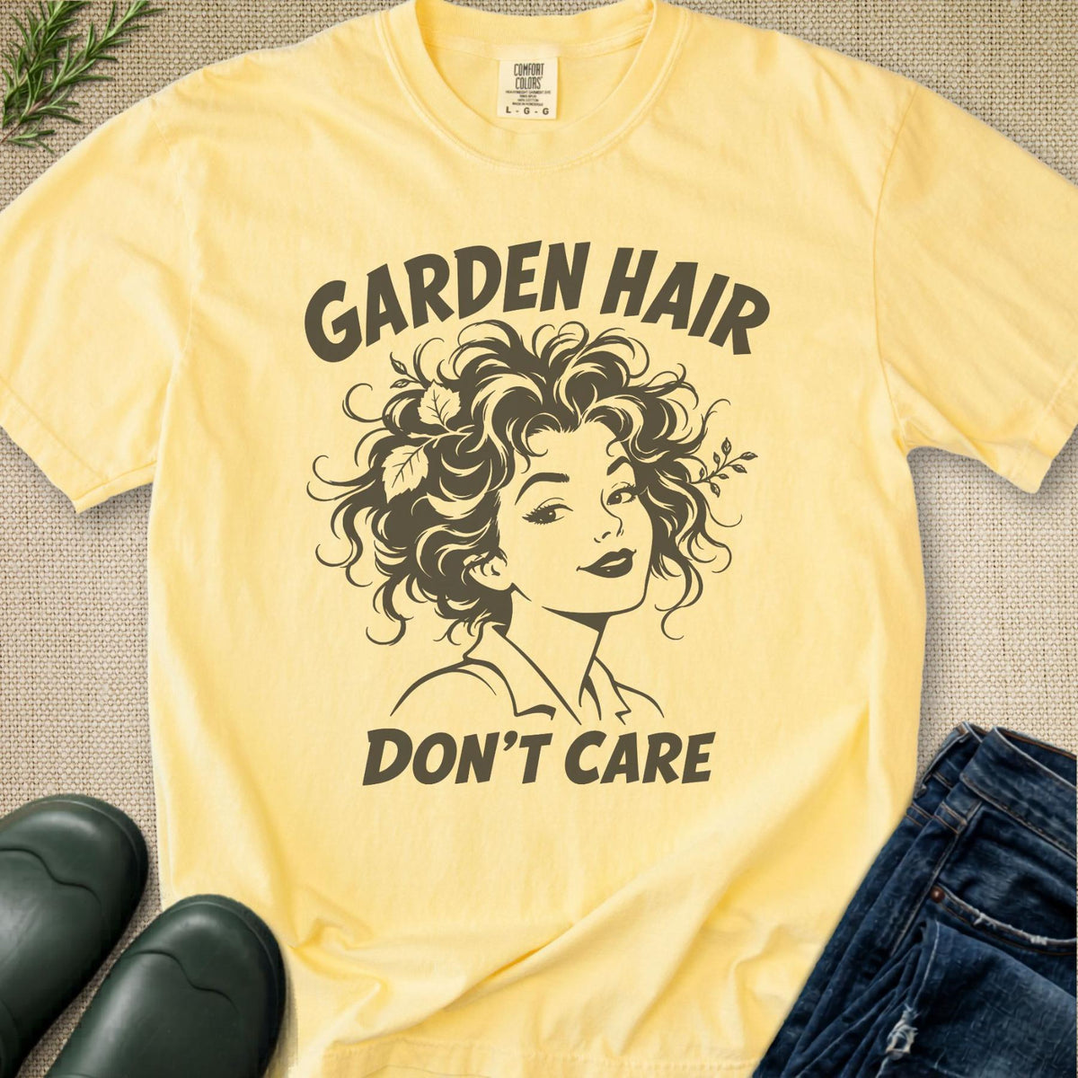 A pale yellow graphic T-shirt laid flat on a textured beige surface, with dark brown text reading 'GARDEN HAIR DON'T CARE' and an illustrated woman with curly hair. Jeans, green rain boots, and a small sprig of greenery are placed around it as styling props.