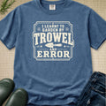 A blue crewneck t-shirt with a white graphic that reads 'I Learnt to Garden by Trowel and Error' with a trowel illustration, styled flat-lay on a burlap background with jeans, boots, and greenery.