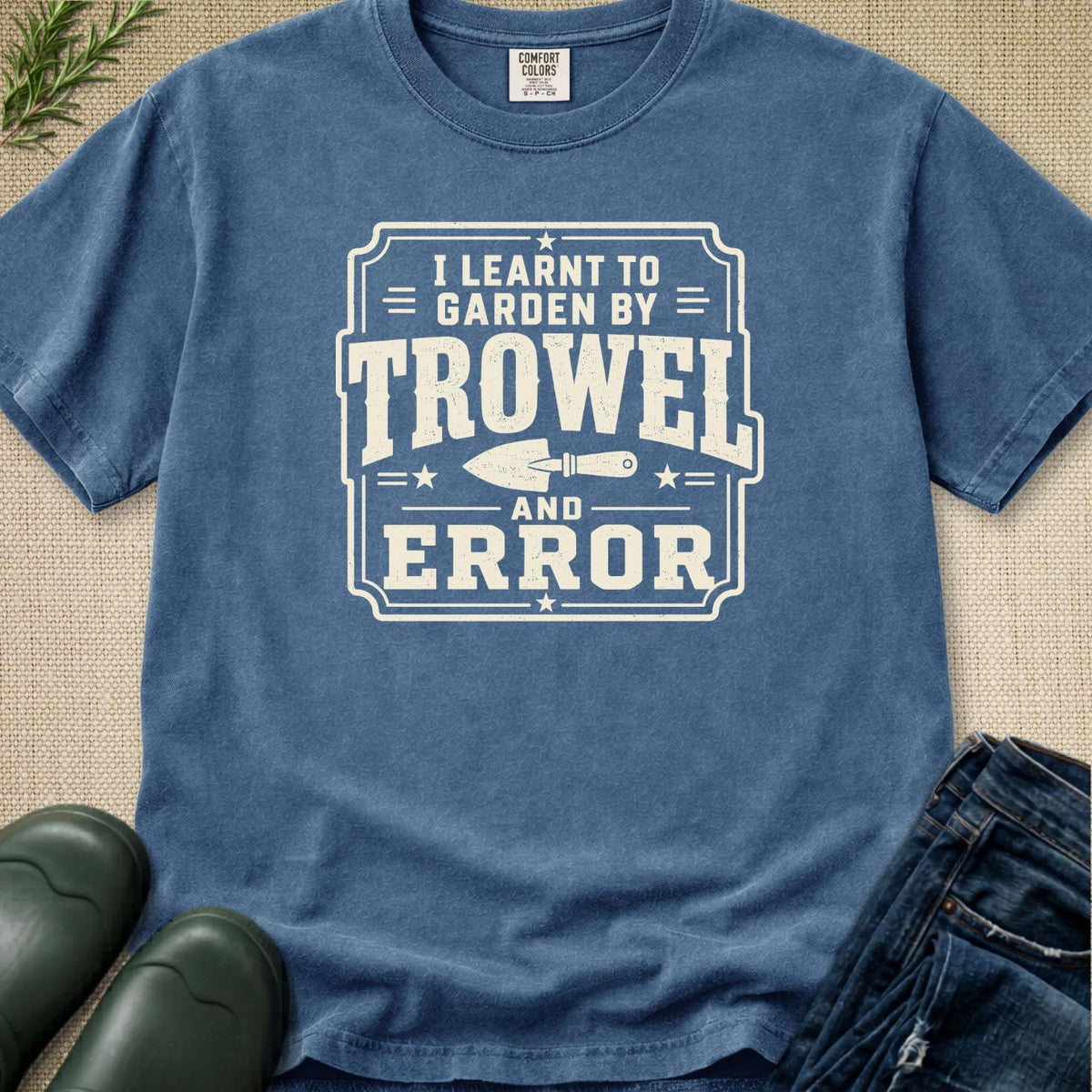 A blue crewneck t-shirt with a white graphic that reads 'I Learnt to Garden by Trowel and Error' with a trowel illustration, styled flat-lay on a burlap background with jeans, boots, and greenery.