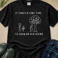 A black short-sleeve graphic T-shirt laid flat on a beige textured surface, with white text reading 'IT TAKES A LONG TIME TO GROW AN OLD FRIEND' and a simple line drawing of two figures and a tree. Partial jeans and dark green boots are visible around the shirt as styling props.