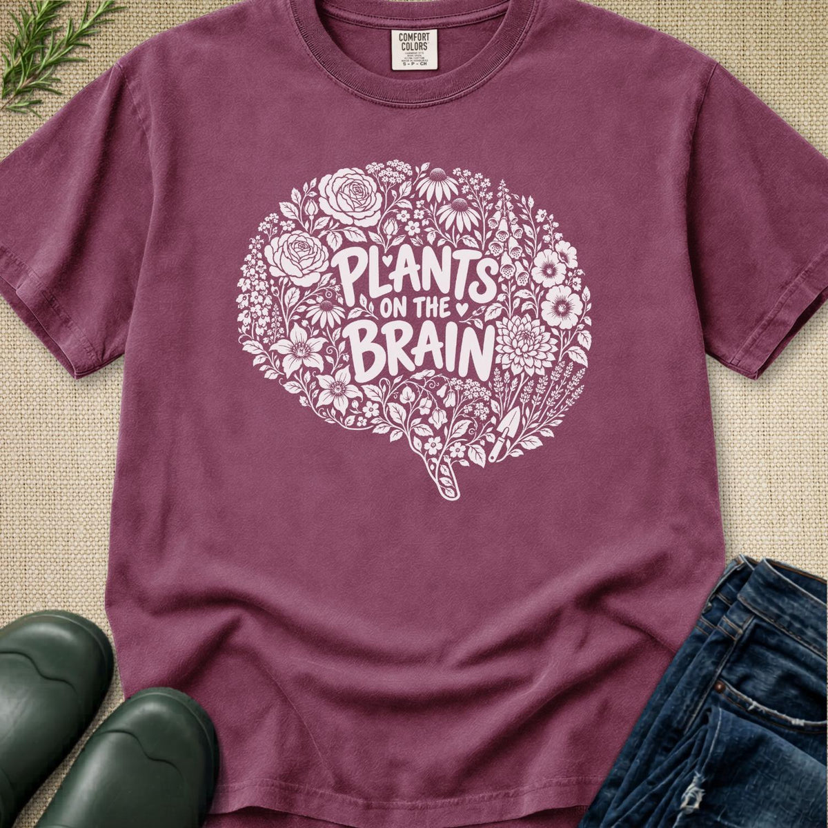 A mauve-purple short-sleeve t-shirt with a white graphic of a brain made of flowers and the text 'PLANTS ON THE BRAIN'. The shirt is styled in a flat-lay with dark green rain boots, blue jeans, and a sprig of greenery on a beige burlap background.