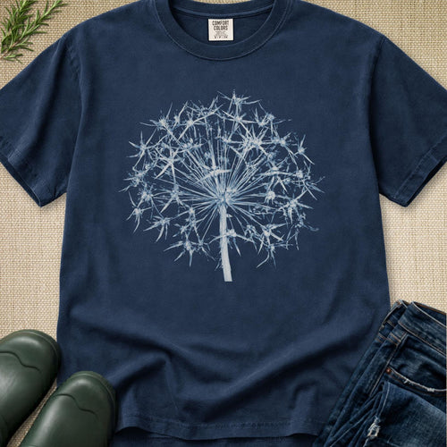 A navy blue crewneck t-shirt featuring a white dandelion graphic on the front, styled in a flat-lay arrangement with dark green rain boots, blue jeans, and a sprig of greenery on a beige textured background.