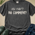 A dark gray graphic t-shirt laid flat on a beige textured background, styled with green boots, blue jeans, and a sprig of greenery. The shirt features the text 'NEW PLANT? NO COMMENT!' in a white, hand-drawn font.