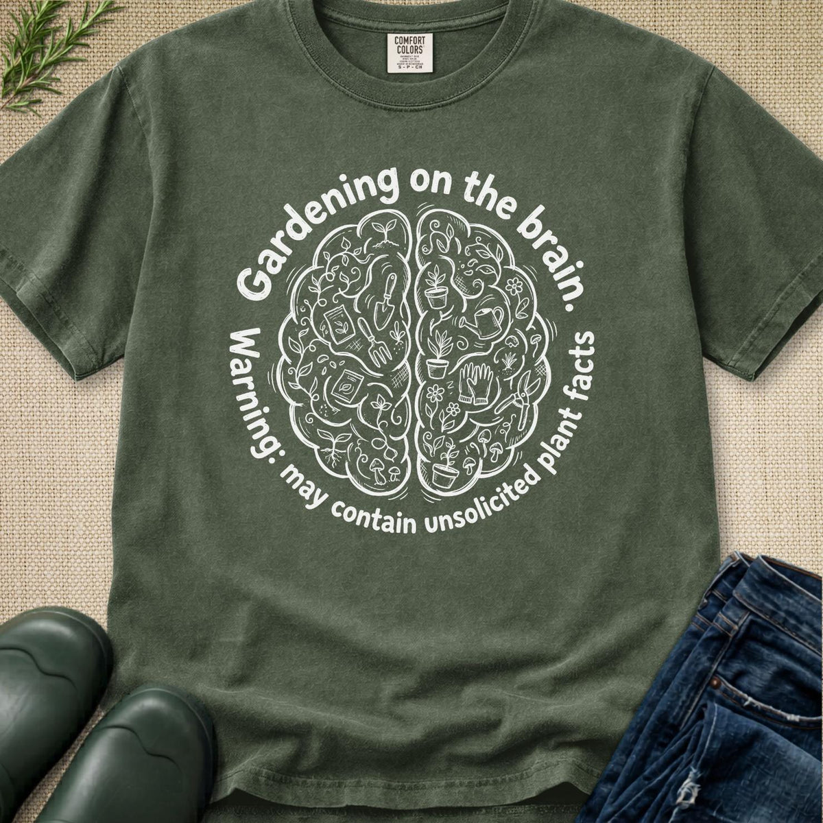 An olive green graphic t-shirt laid flat on a textured beige fabric background, styled with green leaves, black rain boots, and blue jeans. The t-shirt features a white circular graphic of a brain filled with gardening tools and the text 'Gardening on the brain.