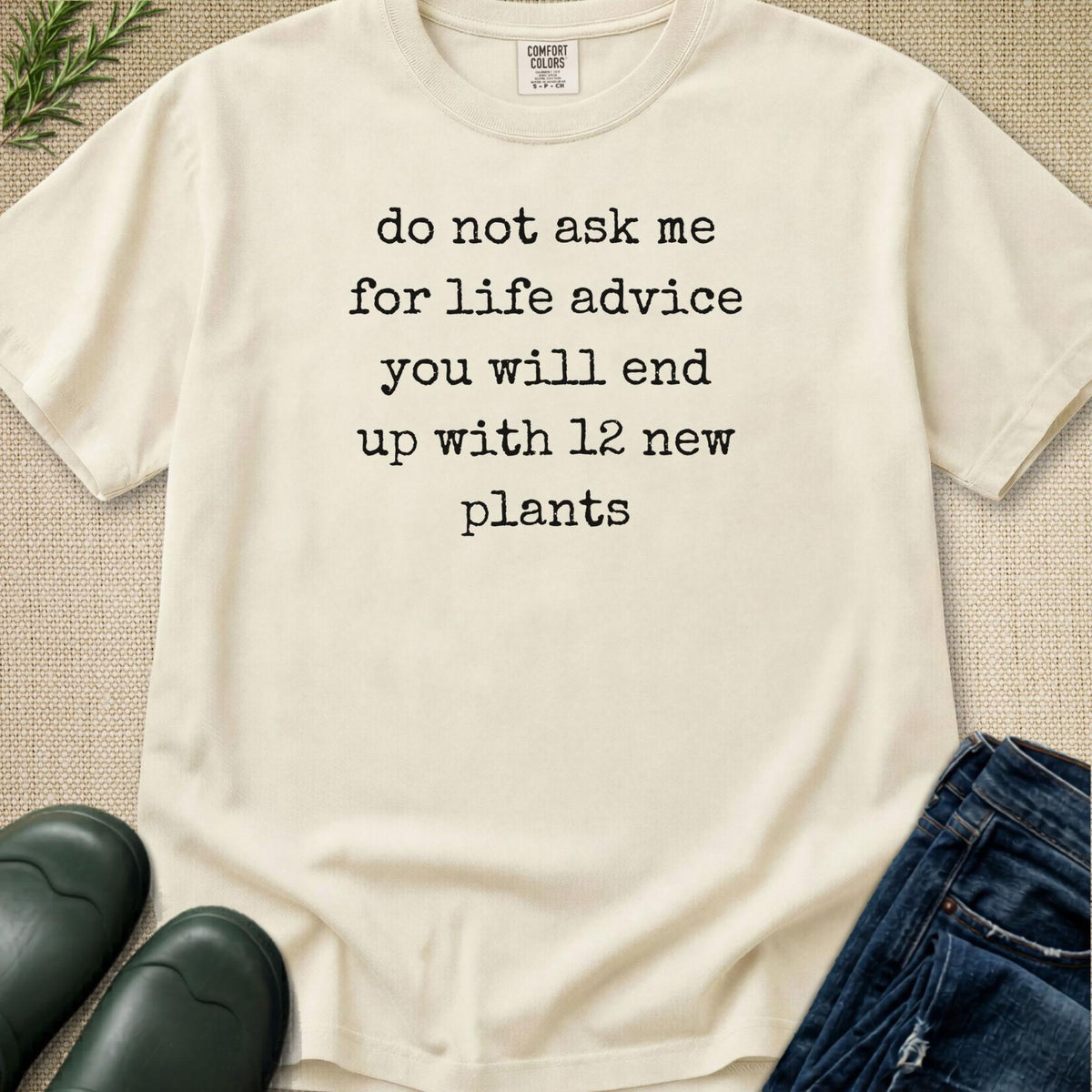 An off-white graphic t-shirt with black text reading 'do not ask me for life advice you will end up with 12 new plants', styled in a flat lay with blue jeans, green rain boots, and a small green plant on a beige textured background.
