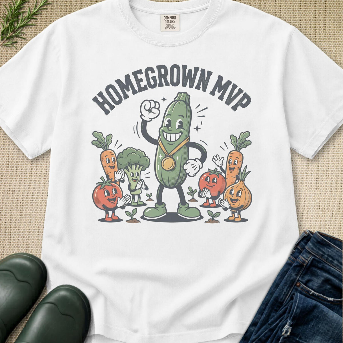 A white short-sleeve graphic T-shirt laid flat on a beige textured surface, with cartoon vegetables and the text 'HOMEGROWN MVP' printed on the front. Jeans and dark green rain boots are partially visible around it as styling props.