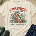 An off-white graphic t-shirt featuring a retro-style illustration of tomatoes, peppers, and asparagus with the text 'NEW JERSEY THE GARDEN STATE' and 'GROW ACCORDINGLY'. The shirt is styled in a flat-lay arrangement with dark green rain boots, blue jeans, and a textured beige fabric background.