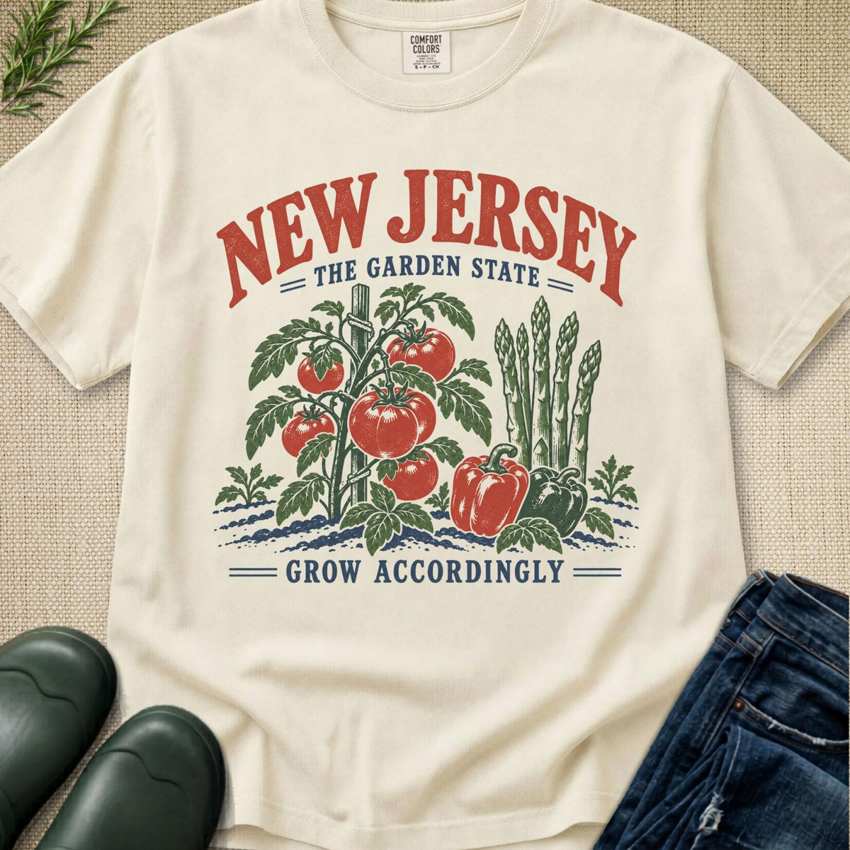 An off-white graphic t-shirt featuring a retro-style illustration of tomatoes, peppers, and asparagus with the text 'NEW JERSEY THE GARDEN STATE' and 'GROW ACCORDINGLY'. The shirt is styled in a flat-lay arrangement with dark green rain boots, blue jeans, and a textured beige fabric background.
