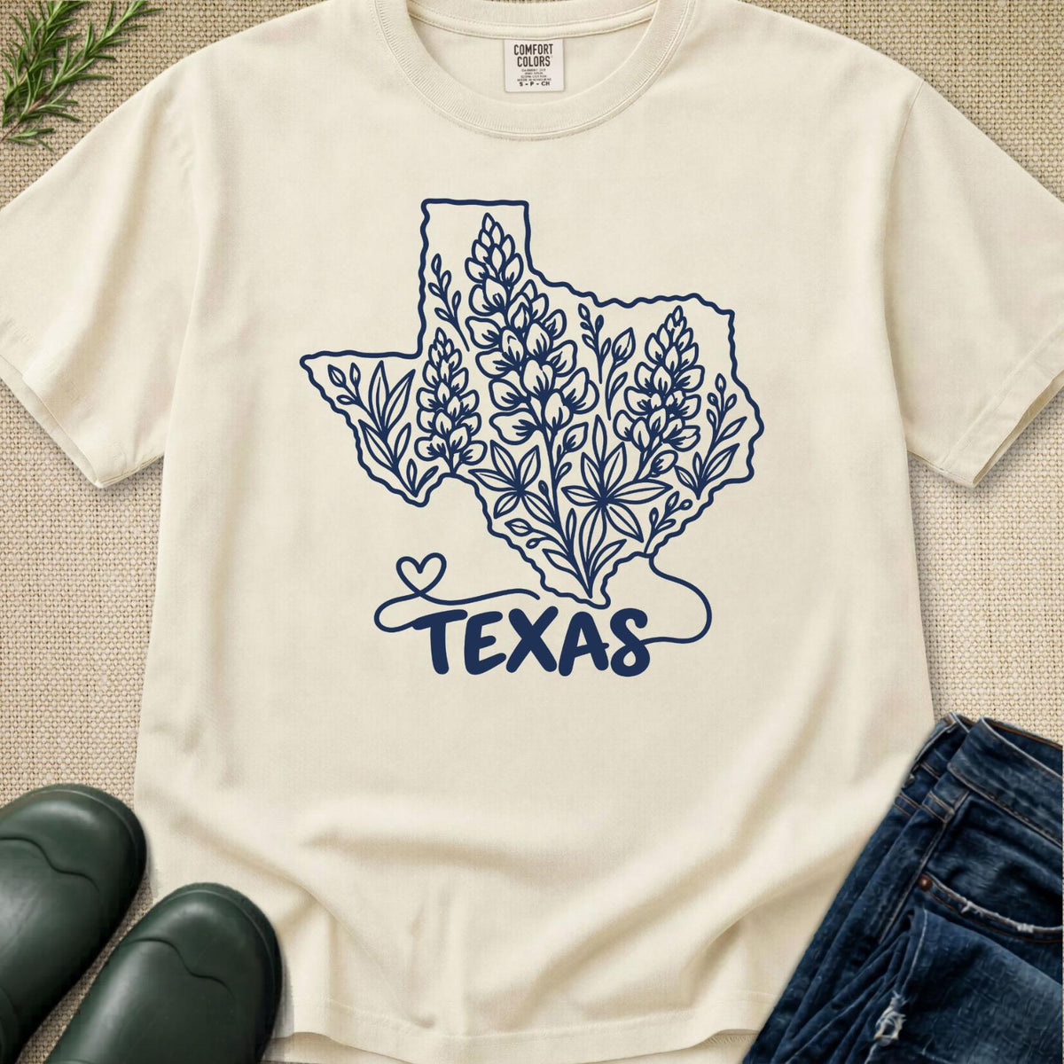 An off-white short-sleeve t-shirt with a navy blue graphic of the state of Texas filled with floral designs and the word 'TEXAS' underneath, styled in a flat lay with jeans, boots, and a small green plant on a textured beige background.