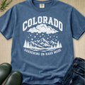 A blue graphic t-shirt laid flat on a burlap background, styled with jeans, green boots, and a sprig of pine. The t-shirt features a white graphic of mountains, trees, rain, and clouds with the text 'COLORADO' and 'GARDENING ON HARD MODE'.