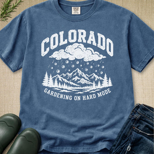 A blue graphic t-shirt laid flat on a burlap background, styled with jeans, green boots, and a sprig of pine. The t-shirt features a white graphic of mountains, trees, rain, and clouds with the text 'COLORADO' and 'GARDENING ON HARD MODE'.