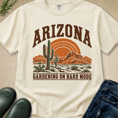An off-white graphic t-shirt featuring a retro desert scene with cacti, mountains, and an orange sun, with the text 'ARIZONA' and 'GARDENING ON HARD MODE'. The shirt is styled in a flat-lay with jeans, boots, and a small plant on a textured beige background.