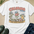 A white graphic t-shirt featuring a retro-style illustration of colorful tomato plants under the word 'COLORADO' and the phrase 'TAKE COVER EVERYONE'. The shirt is styled in a flat-lay arrangement on a beige textured background with dark green rain boots and blue jeans partially visible.