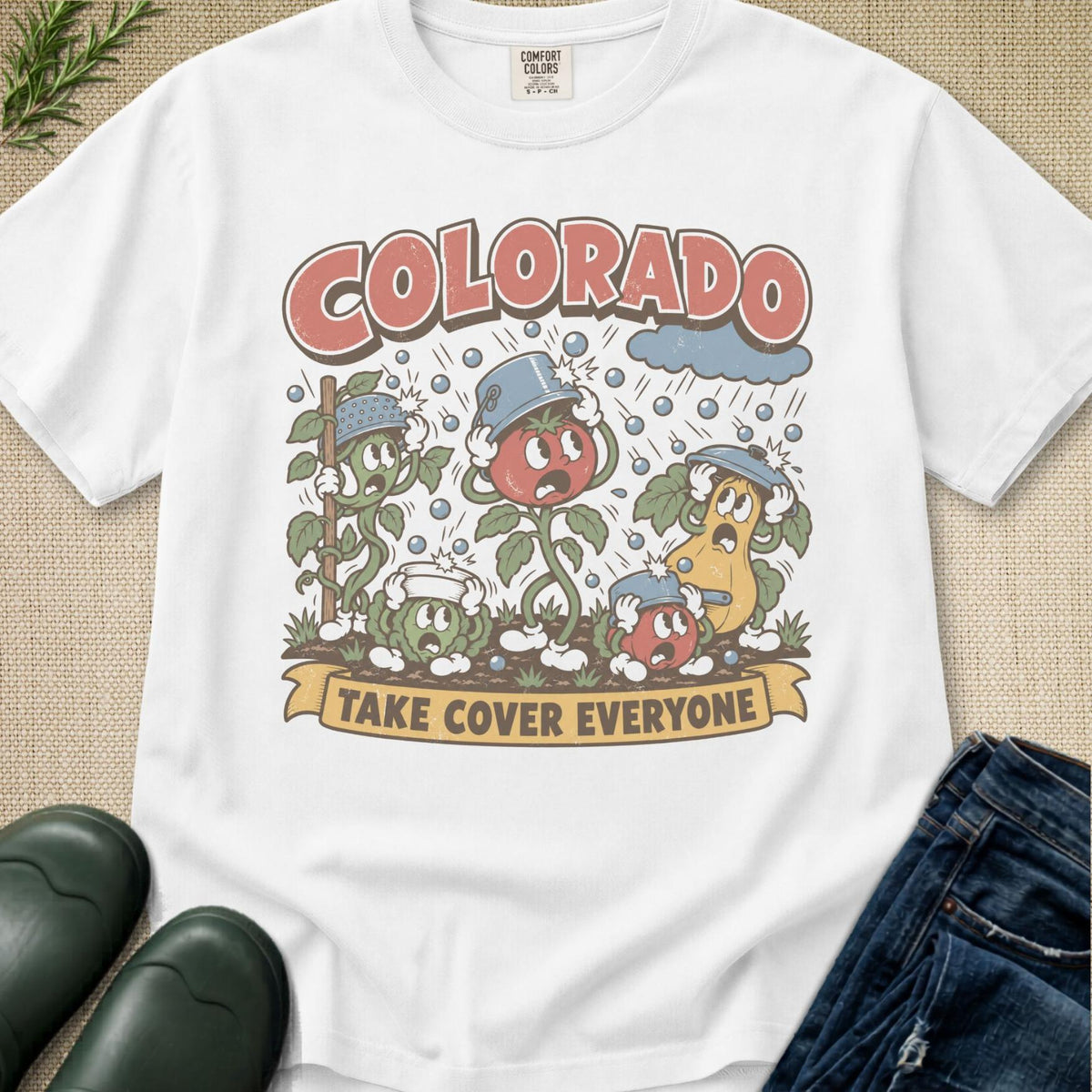 A white graphic t-shirt featuring a retro-style illustration of colorful tomato plants under the word 'COLORADO' and the phrase 'TAKE COVER EVERYONE'. The shirt is styled in a flat-lay arrangement on a beige textured background with dark green rain boots and blue jeans partially visible.