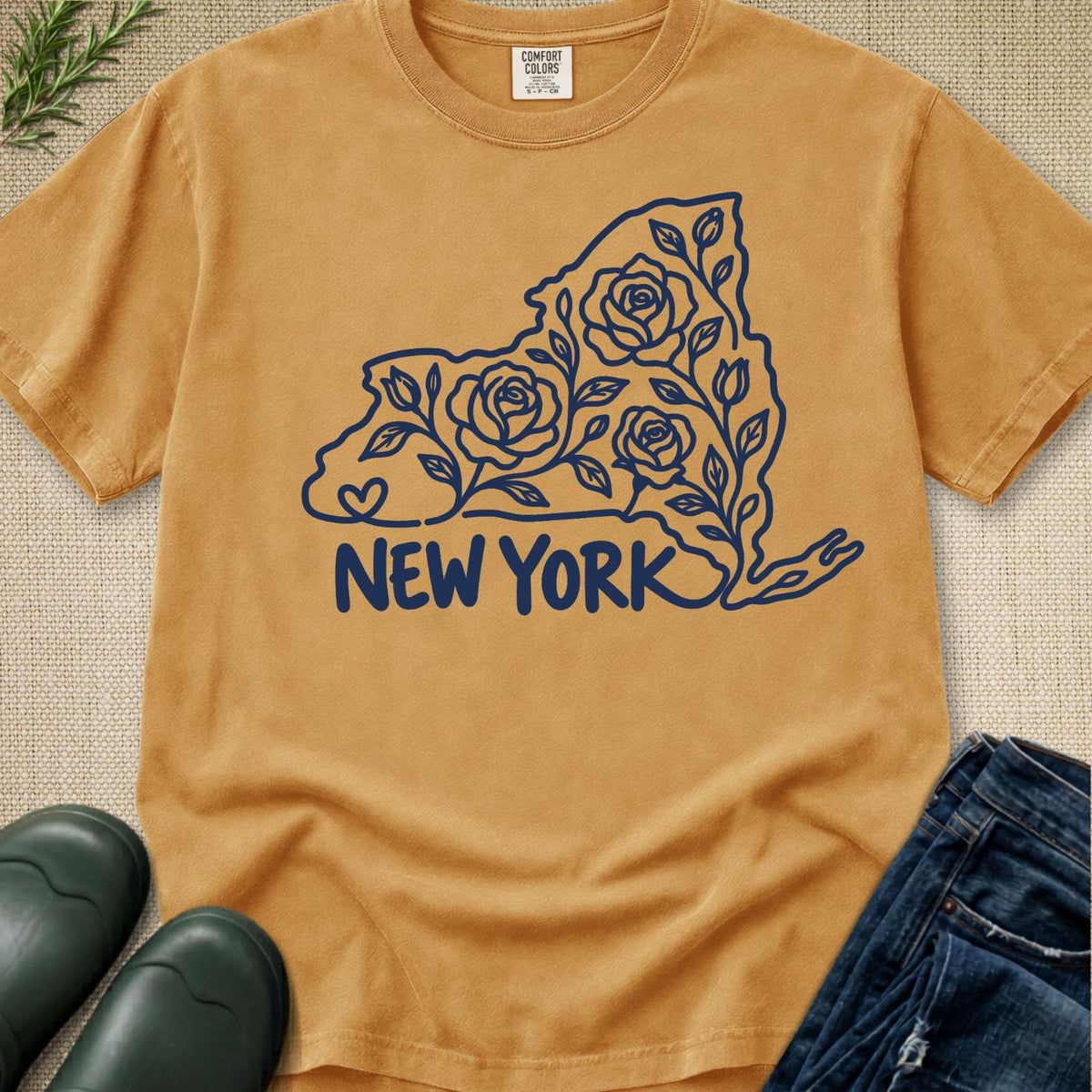 A tan graphic t-shirt featuring a dark blue line art design of the state of New York filled with roses and the text 'NEW YORK' underneath. The shirt is styled in a flat-lay arrangement with dark green rain boots, blue jeans, and a sprig of greenery on a textured beige background.