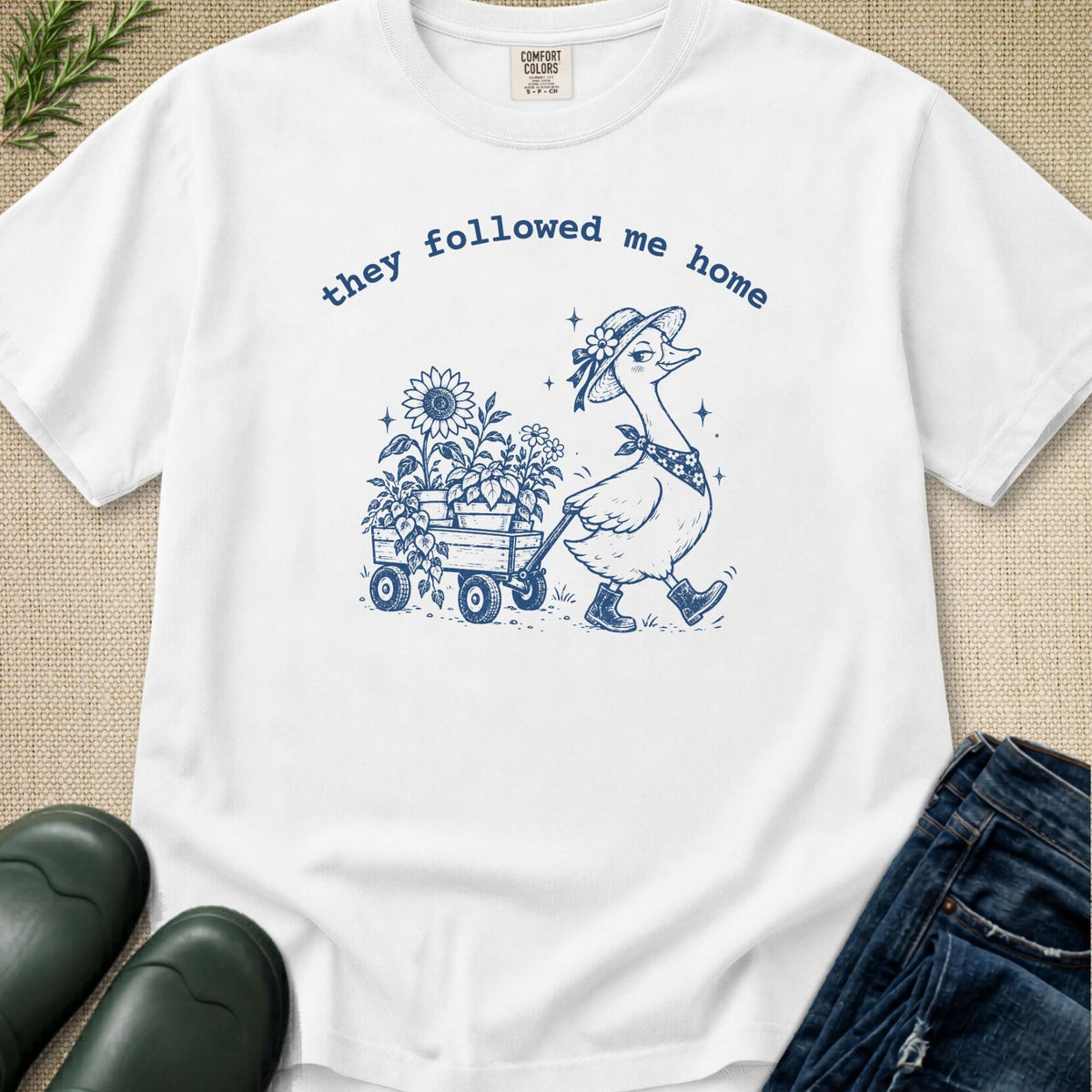 A white short-sleeve graphic T-shirt laid flat on a beige textured surface, with blue text reading 'they followed me home' and a blue line drawing of a goose pulling flowers in a wagon. Jeans, green boots, and a small plant are partially visible around it as styling props.
