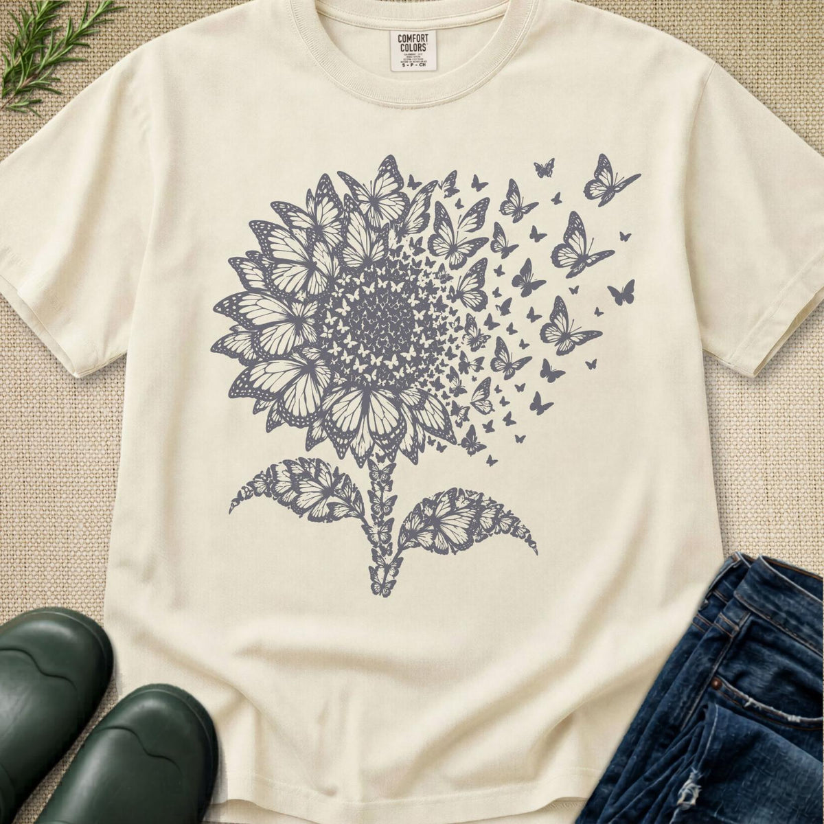 An off-white short-sleeve t-shirt with a dark gray graphic of a sunflower turning into butterflies, styled in a flat-lay with jeans, rain boots, and greenery.