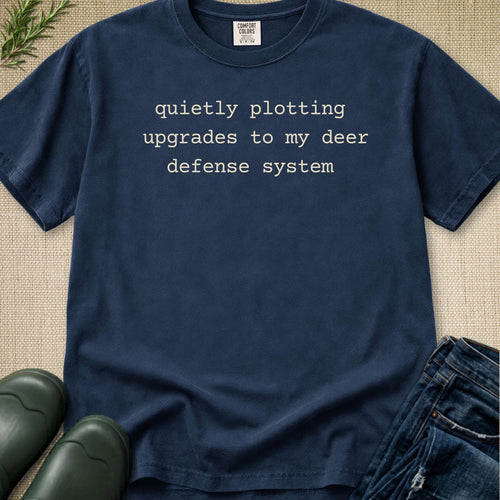 A navy blue crewneck t-shirt with the text 'quietly plotting upgrades to my deer defense system' printed in a light beige typewriter font. The shirt is styled in a flat-lay arrangement on a textured beige burlap background, accompanied by dark green boots and blue jeans.