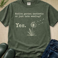 An olive green graphic t-shirt laid flat on a textured beige fabric background, styled with dark green rain boots, blue jeans, and a sprig of greenery. The t-shirt features a white graphic of a dandelion and the text 'Native garden aesthetic or just hate weeding? Yes.