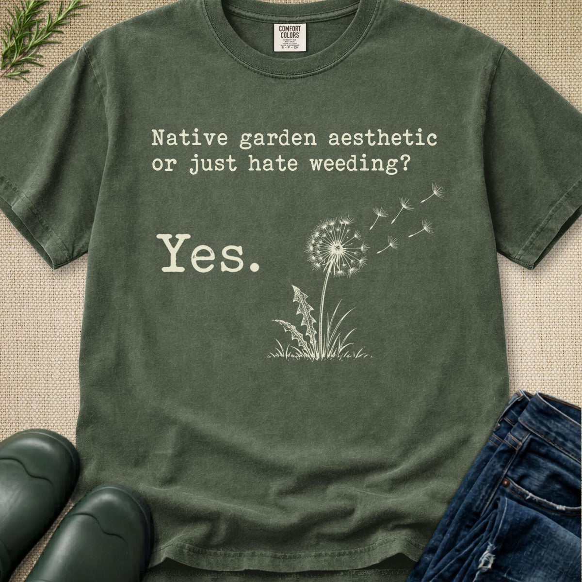 An olive green graphic t-shirt laid flat on a textured beige fabric background, styled with dark green rain boots, blue jeans, and a sprig of greenery. The t-shirt features a white graphic of a dandelion and the text 'Native garden aesthetic or just hate weeding? Yes.