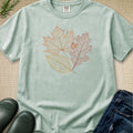 A mint green crewneck t-shirt with a graphic of three autumn leaves (maple, oak, and bittersweet) in shades of yellow, peach, and pink. The shirt is styled in a flat-lay arrangement on a beige woven mat, accompanied by dark green rain boots, dark blue jeans, and a sprig of green pine needles.
