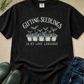A black short-sleeve graphic T-shirt laid flat on a beige textured surface, with white text reading 'GIFTING SEEDLINGS IS MY LOVE LANGUAGE' and an illustration of five seedling plants in pots. Partial jeans, green rubber boots, and a small plant are visible around the shirt as styling props.