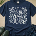 A navy blue graphic t-shirt with a white hand-lettered design that reads 'DIRT ON MY HANDS, PEACE IN MY HEART' with illustrations of flowers, a wheelbarrow, and a gardening tool. The shirt is styled in a flat-lay arrangement on a beige textured background with green rain boots and blue jeans partially visible.