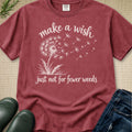 A maroon short-sleeve graphic T-shirt laid flat on a textured beige surface, with white text reading 'make a wish just not for fewer weeds' and a dandelion illustration. Jeans, green rain boots, and a small green sprig are styled around it.