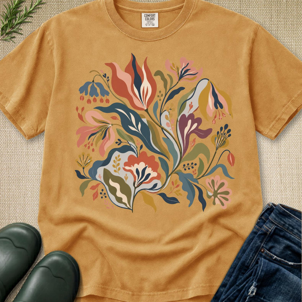 A mustard yellow short-sleeve t-shirt with a colorful botanical floral print, styled in a flat-lay with dark green rain boots, blue jeans, and a sprig of greenery on a textured beige background.